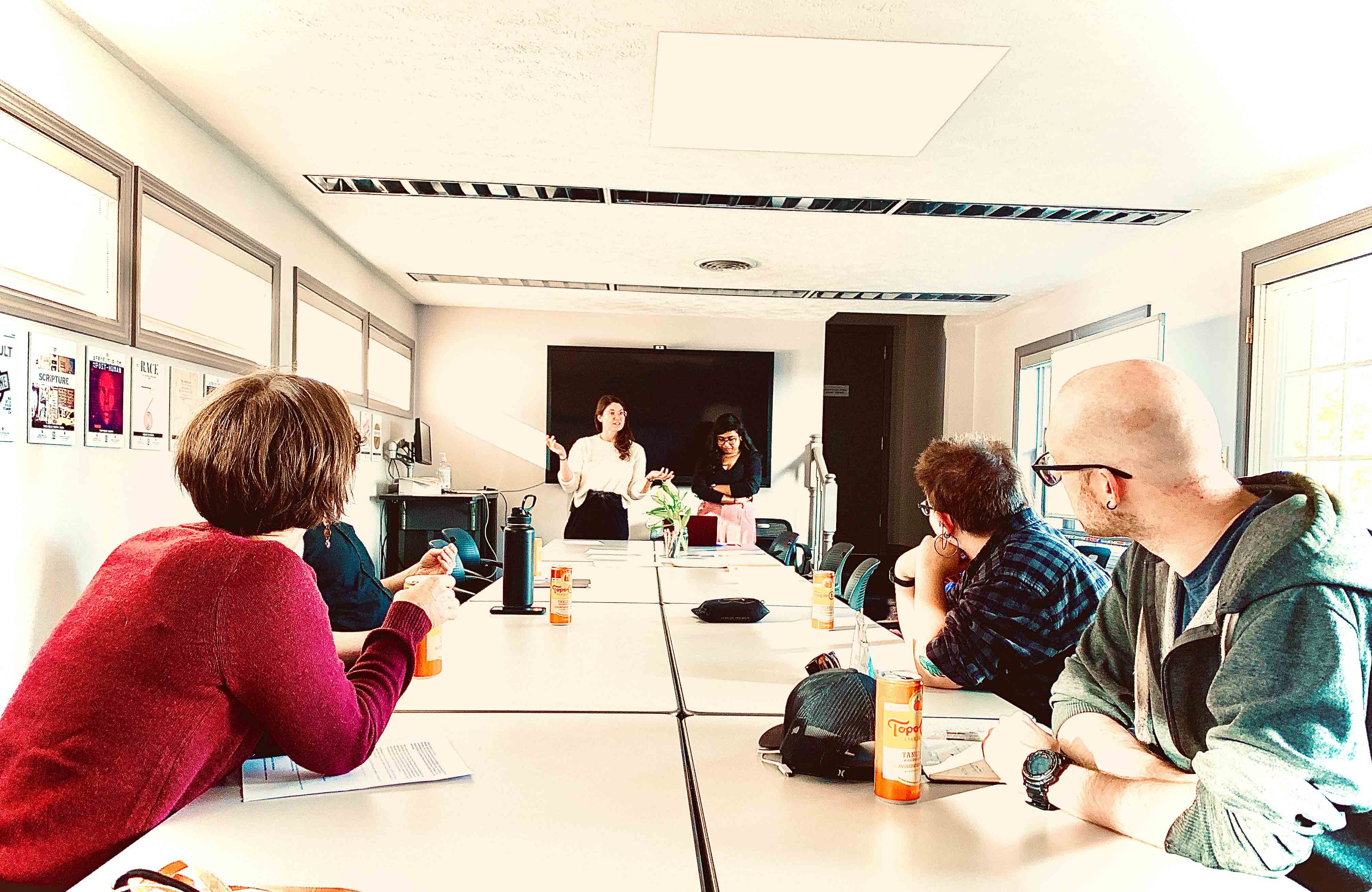 Four people convened around a conference table while two people present to them from the front of the room.