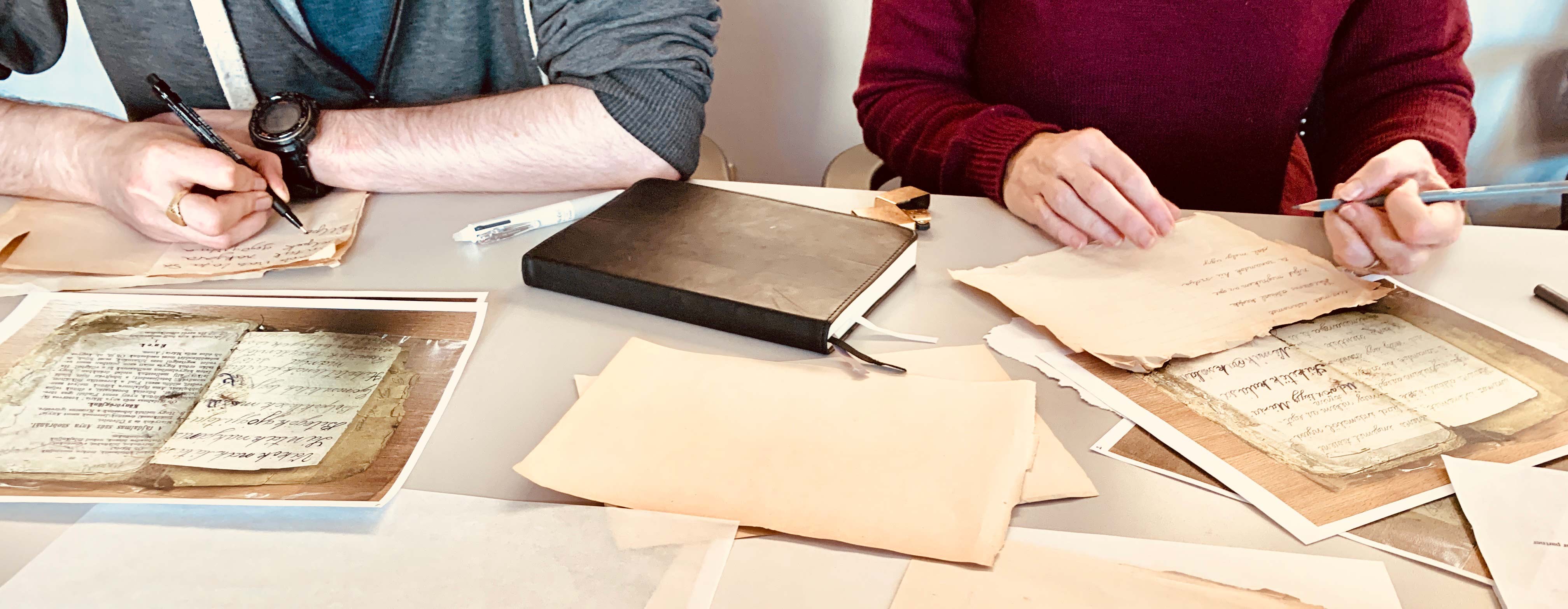 A table covered in papers and notebooks, two people sitting at it, cropped to show their hands as they transcribe.